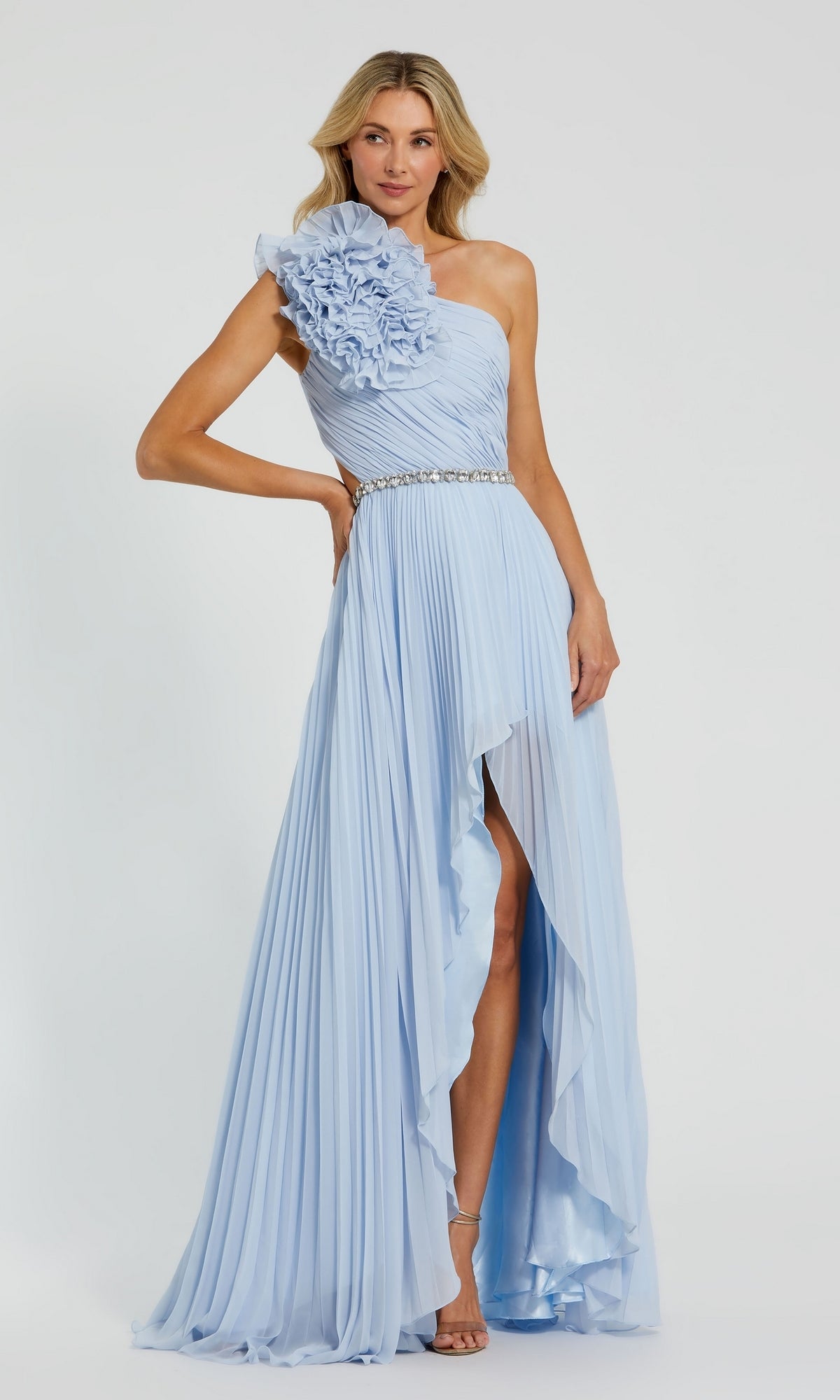 A woman wears the Long Formal Dress 49687 by Mac Duggal, a powder blue one-shoulder A-line gown with pleats, a large shoulder floral detail, and a beaded belt, standing against a plain background.
